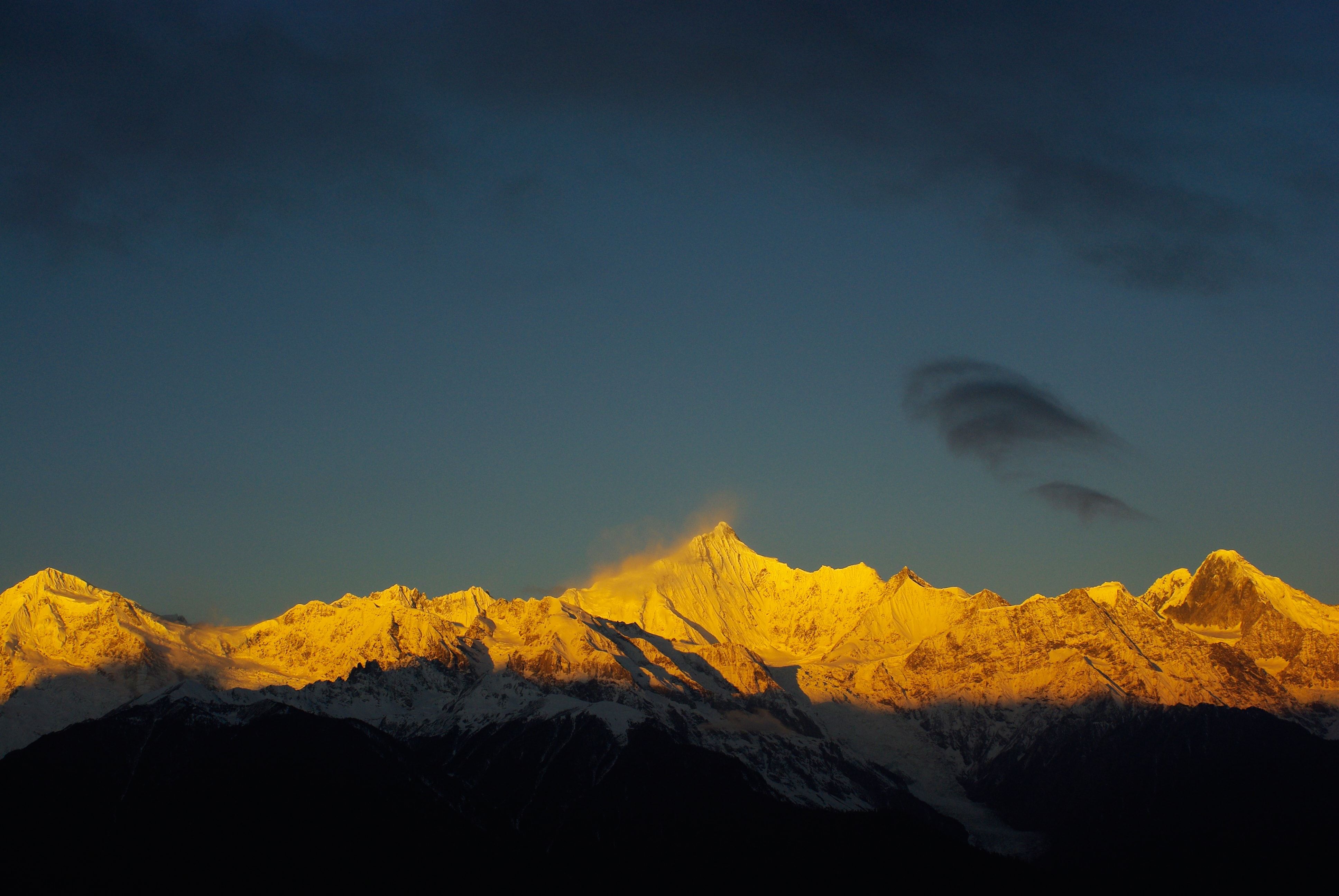 Kawagebo’s summit lit gold at sunrise, snow blowing off the top, under a dark sky