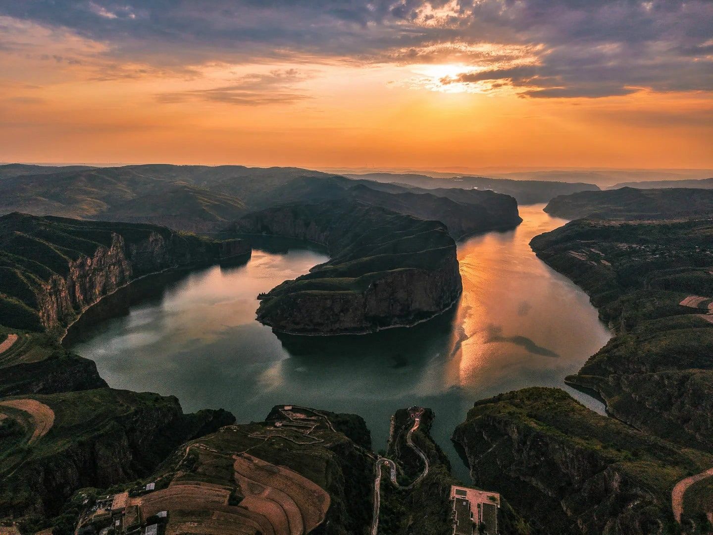 The green Yellow River bending around a cliff-edged spur at sunset, seen from the air, with terraced fields and a winding road below