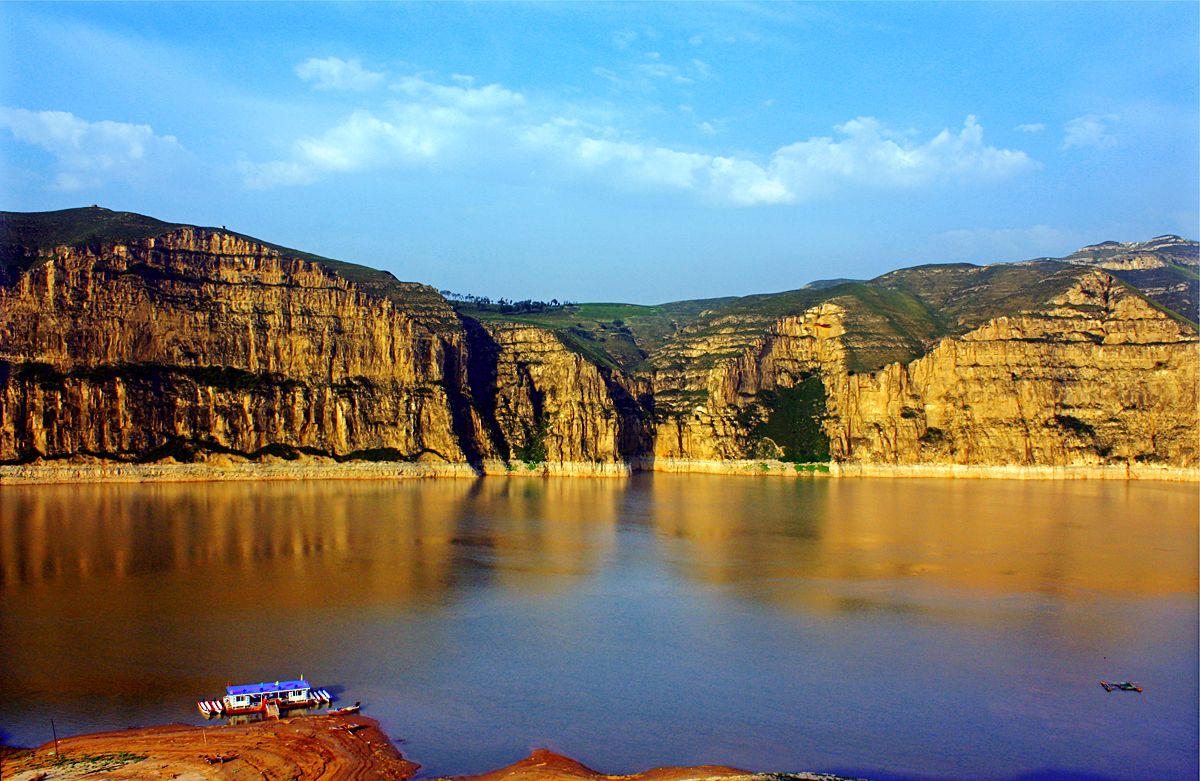 Sheer golden cliffs above the still Yellow River at Laoniuwan, a side canyon opening between them and a small ferry on the shore