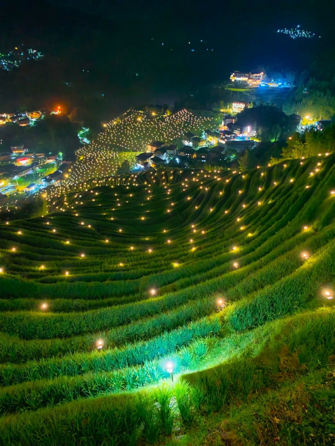 Terrassen bei Nacht, gesäumt von kleinen Solarlampen, darüber ein leuchtendes Dorf