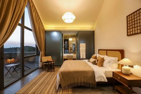 A room at Yellow River Qiankun B&B: the bed beside a glass wall onto a terrace and the hills at dusk