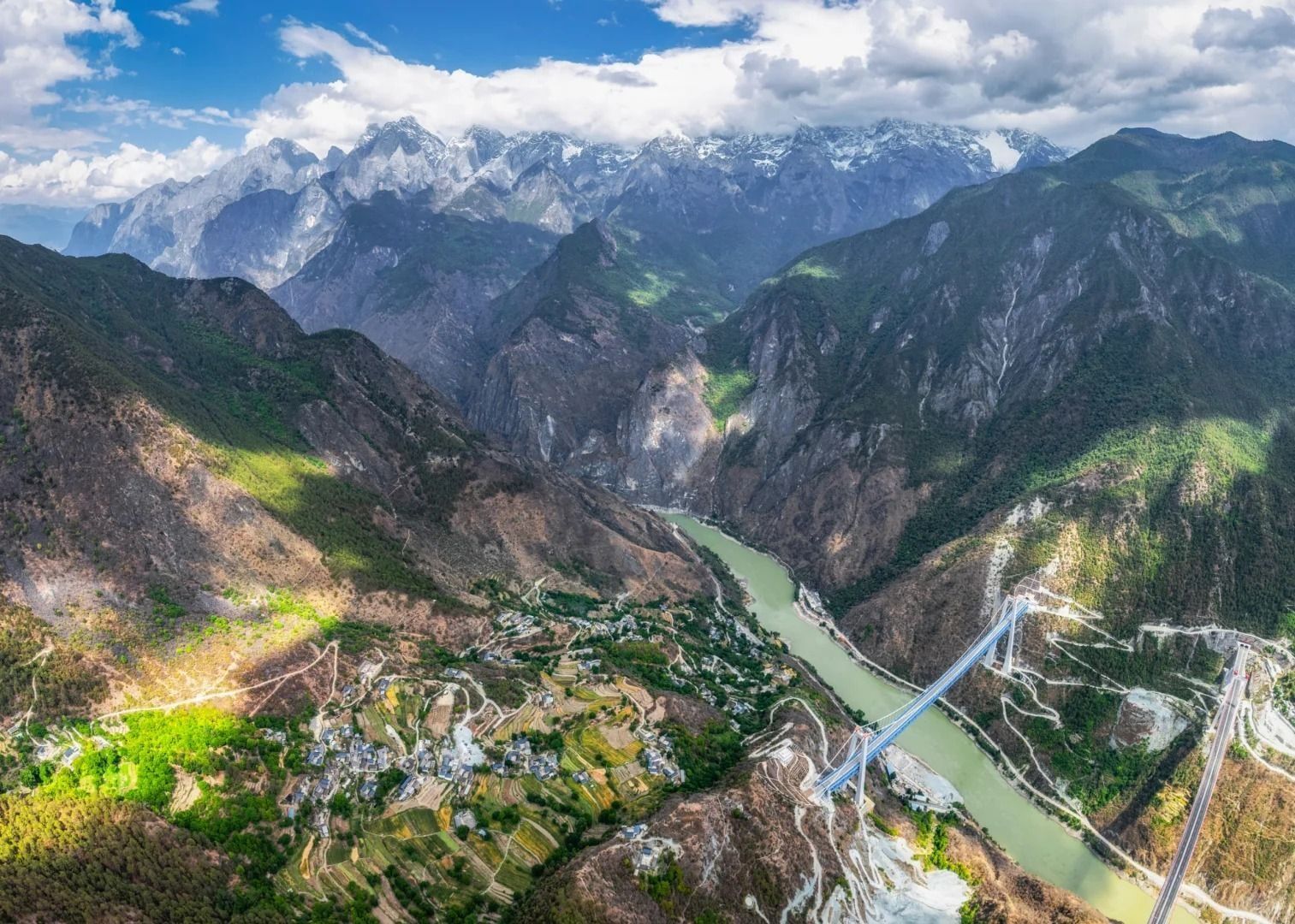 Les gorges du Saut du Tigre vues du ciel : un village au milieu des cultures en terrasses, le Jinsha vert sous un pont suspendu, des sommets gris déchiquetés sous les nuages blancs