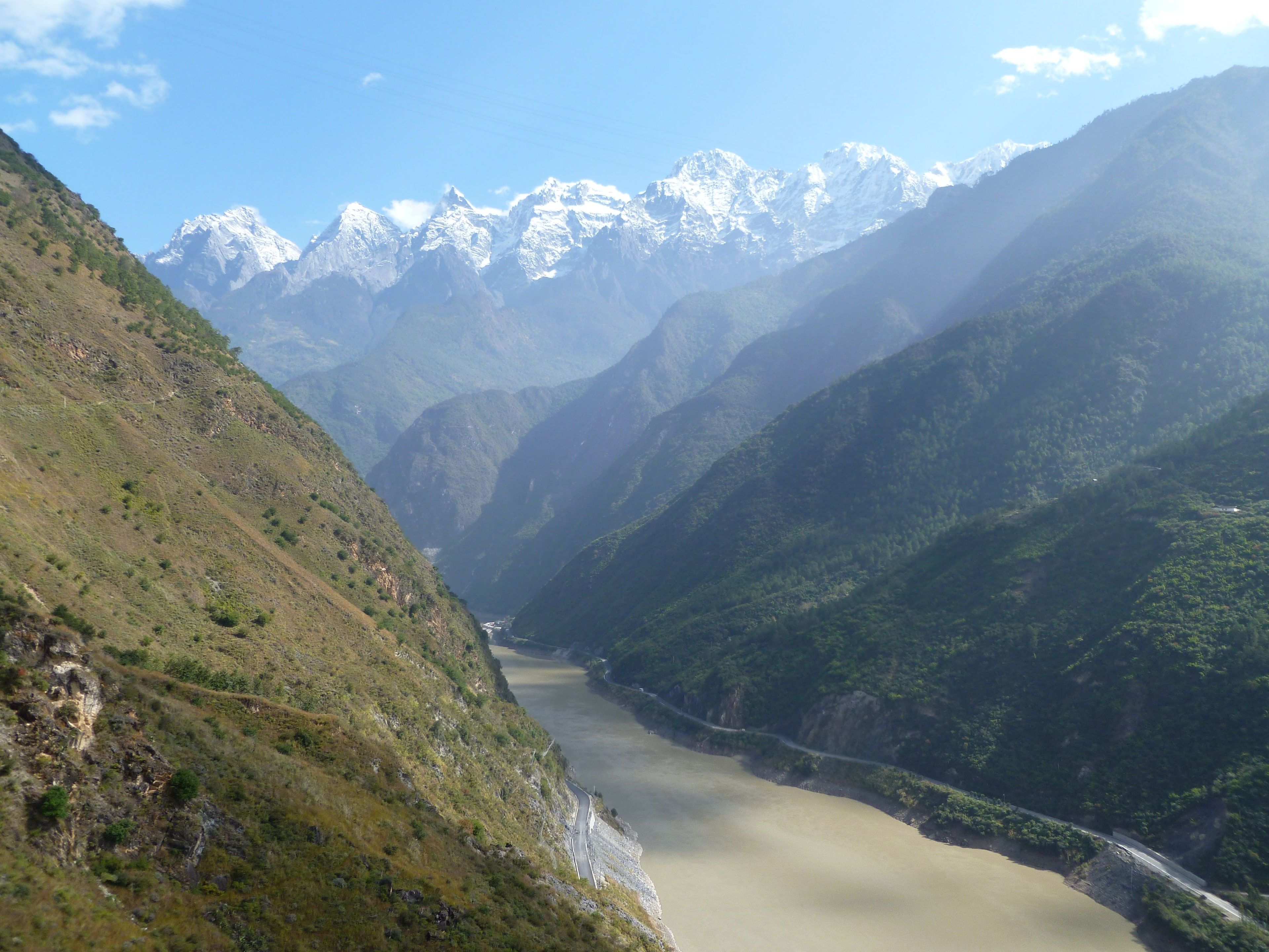 The silt-brown Jinsha River and the road along its bank deep in Tiger Leaping Gorge, the snow peaks of Jade Dragon Snow Mountain at the head of the valley