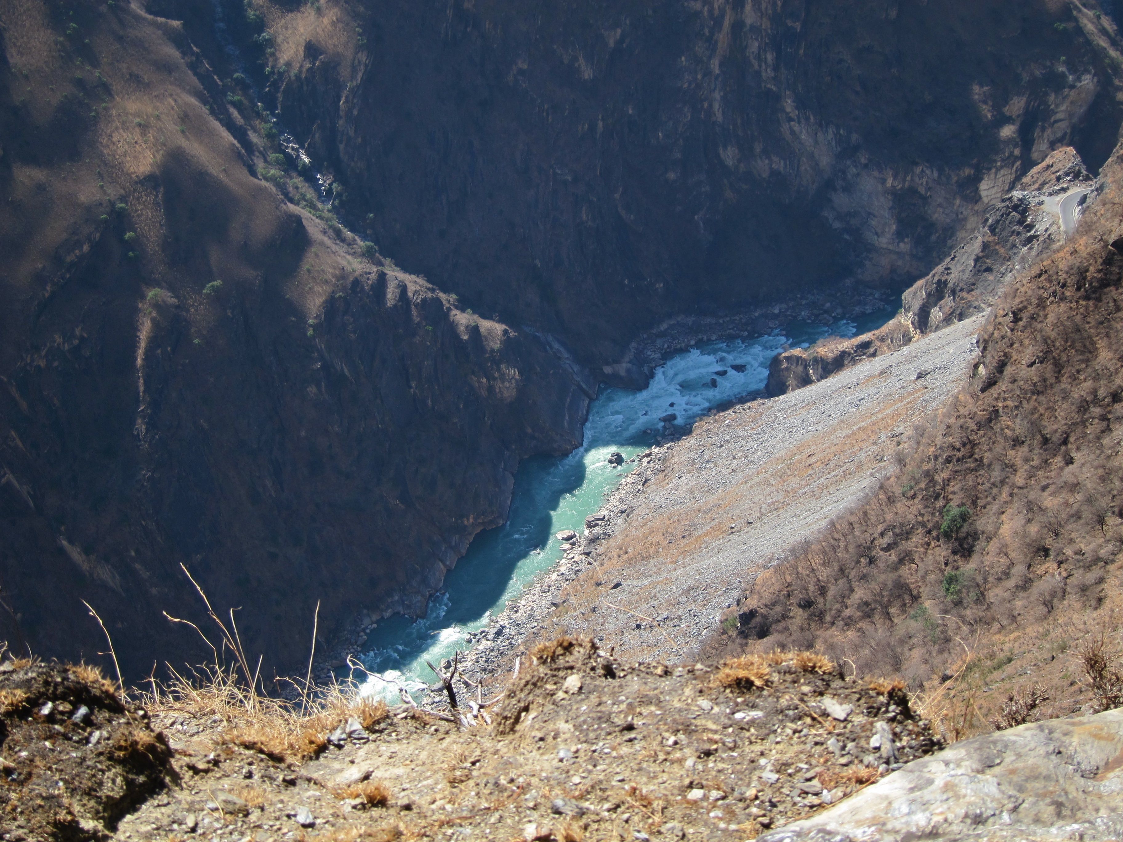 The turquoise Jinsha River foaming through the gorge far below, a grey scree slope beside it, seen from a dry hillside high above