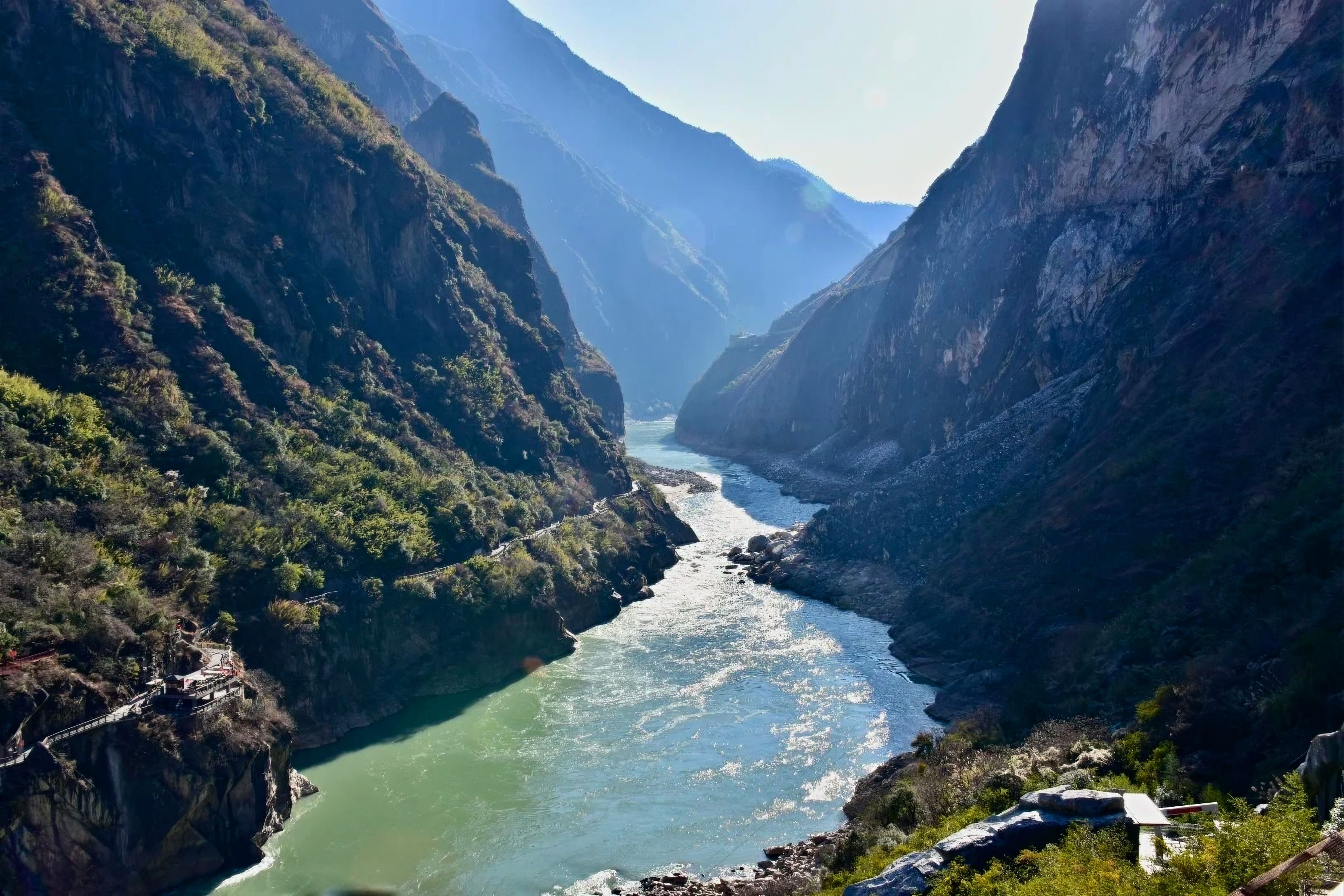 The green Jinsha River running between sheer walls in Tiger Leaping Gorge, a cliffside path on the left, in hazy backlight