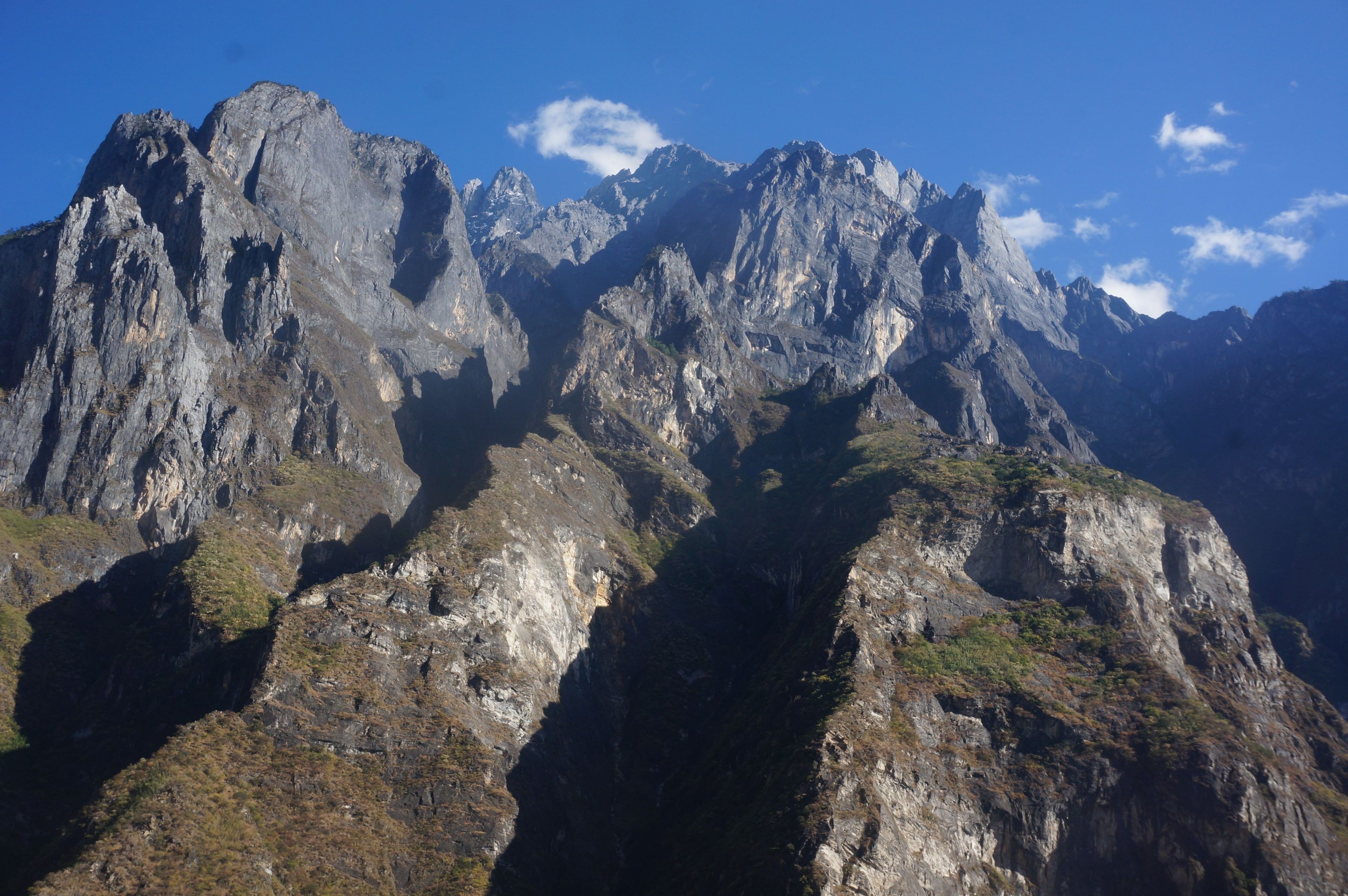 Grey rock peaks and sheer cliffs above Tiger Leaping Gorge in sunshine, small white clouds in a deep blue sky