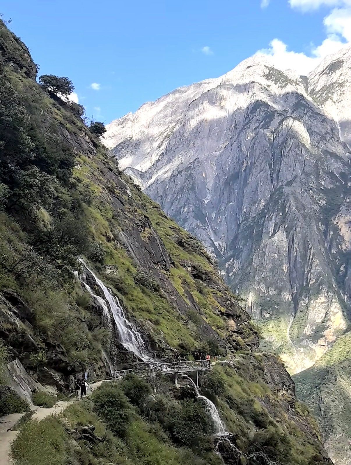 Des randonneurs sur le sentier d’altitude sous la cascade de Guanyin et sa passerelle en bois, face aux parois nues du mont Yulong