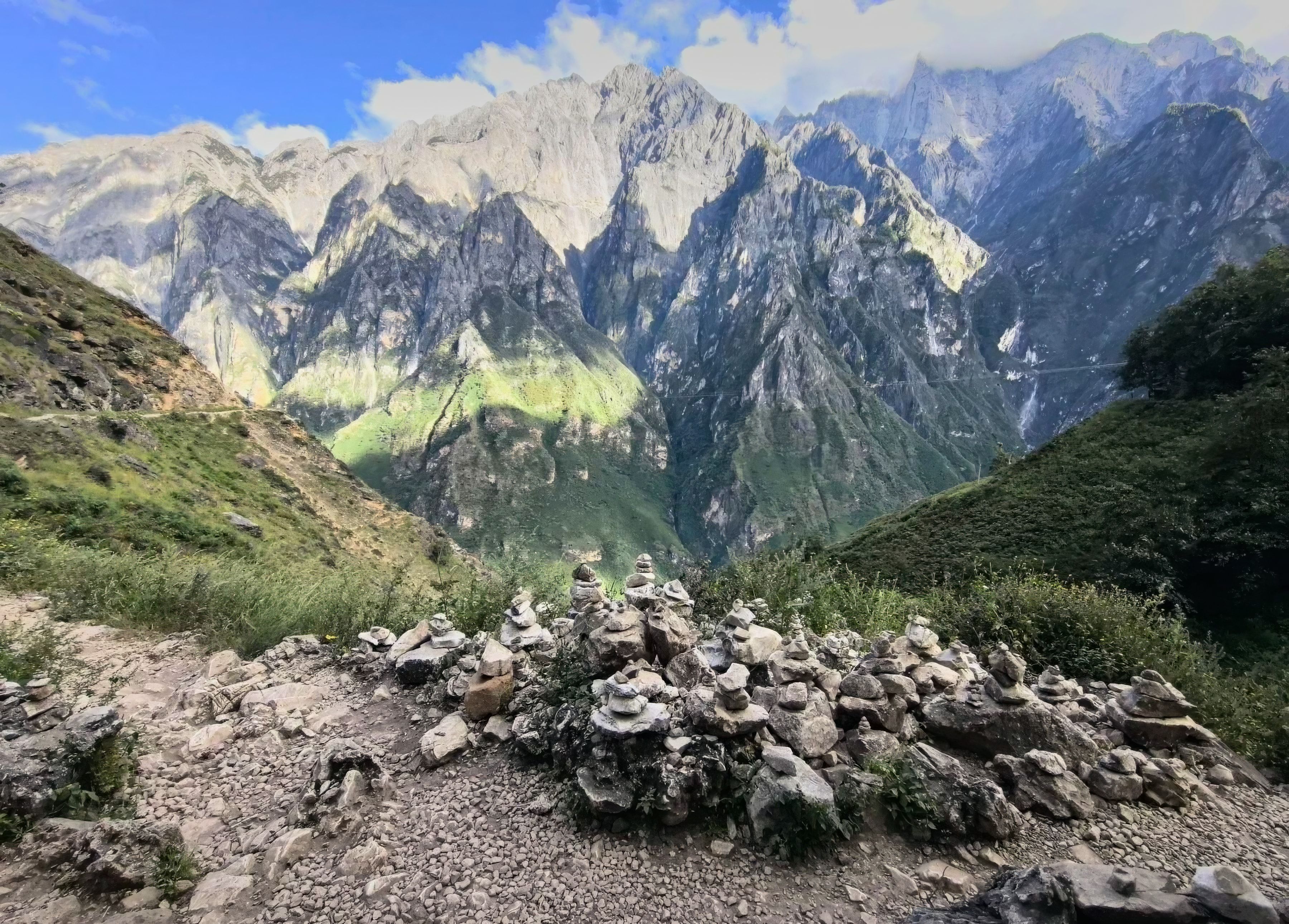 Little stone cairns (mani piles) on the high trail, facing the grey cliffs of Jade Dragon Snow Mountain across the gorge under a blue sky with white cloud