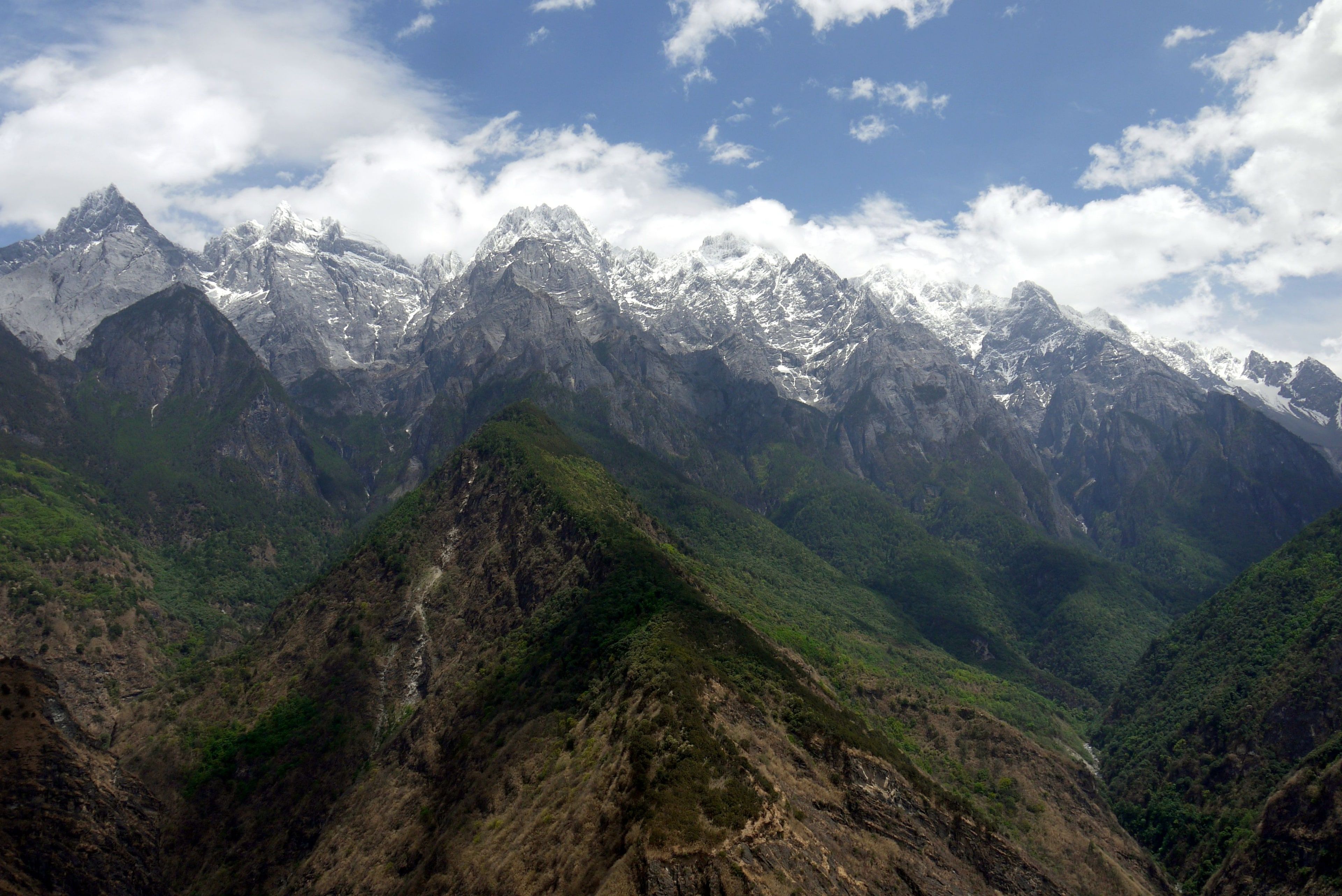 A long ridge of snow peaks above green, forested slopes, white cloud drifting across a blue sky