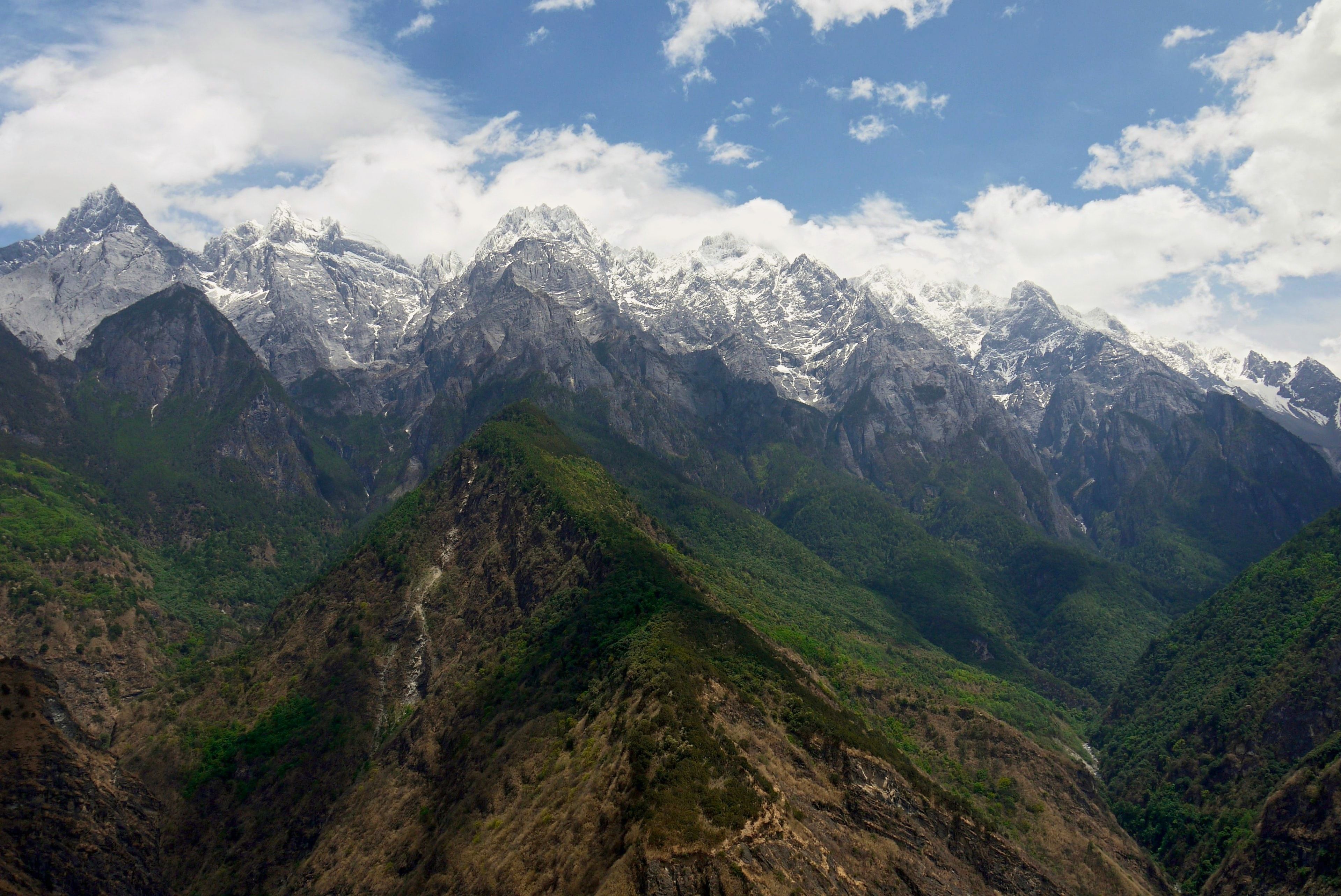 A long ridge of snow peaks above green, forested slopes, white cloud drifting across a blue sky