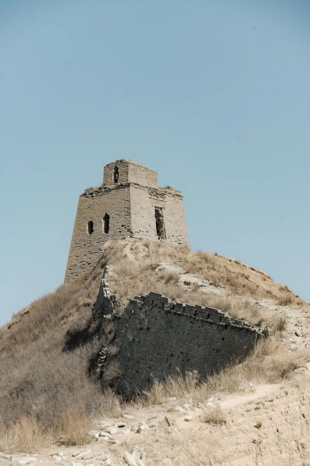 A ruined brick watchtower on a grassy hilltop, a broken stretch of brick wall below it