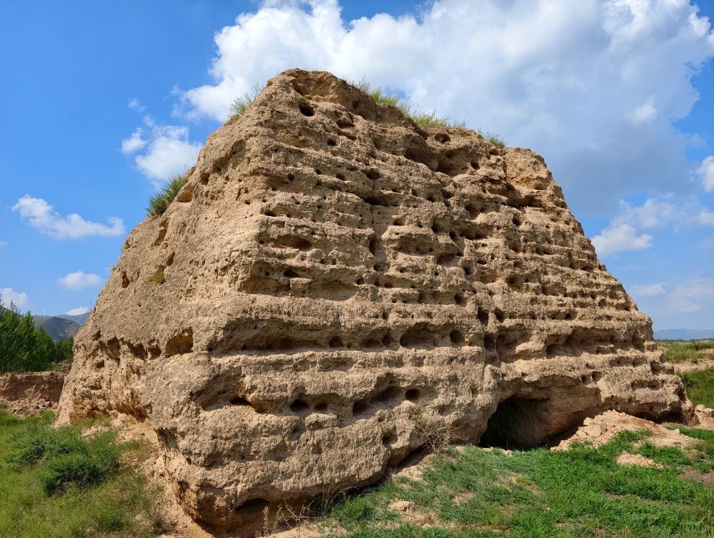 A weathered rammed-earth watchtower pitted with holes, under white clouds and a blue sky