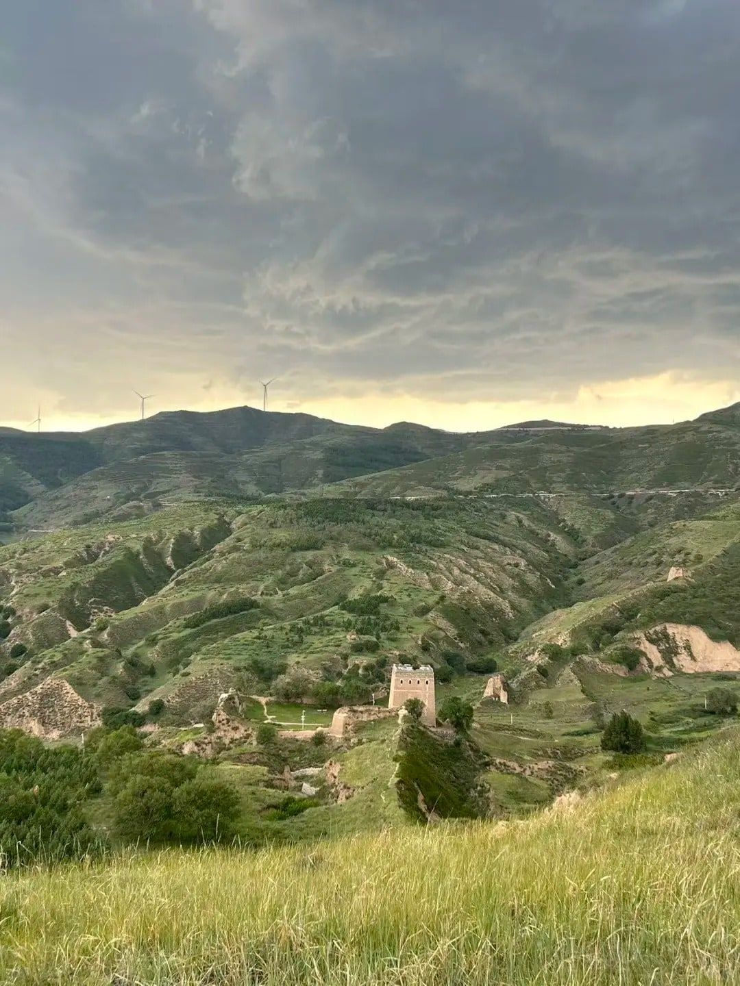 A square brick watchtower in green loess hills under storm clouds, wind turbines on the skyline