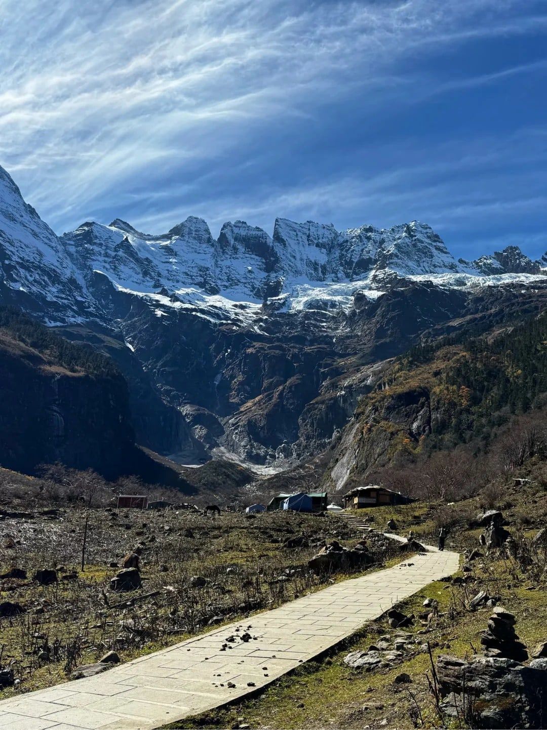 Ein Plattenweg führt zu Hütten unter einer Reihe verschneiter Felstürme und einem Gletscher, unter tiefblauem Himmel