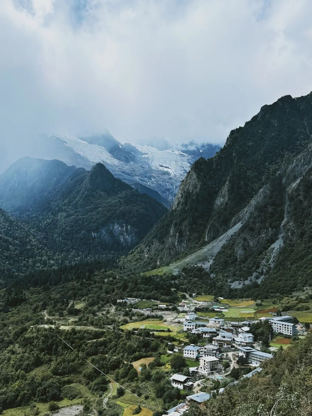 Die weißen Gästehäuser und grünen Felder von Yubeng unten im Tal, dahinter Gletscher und Gipfel unter Wolken