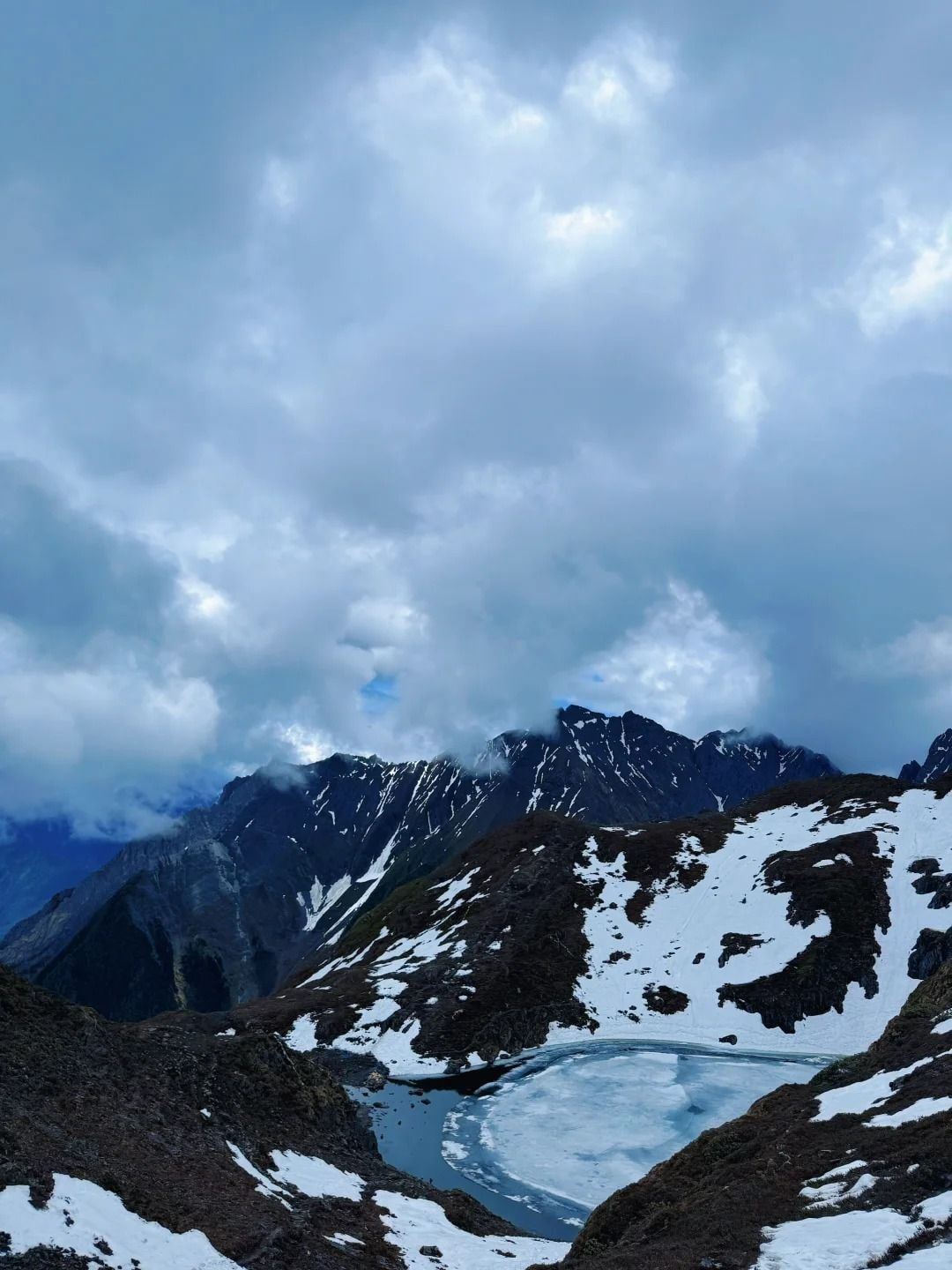 Ein halb zugefrorener See in einer Mulde zwischen Schneeflecken und dunklen Graten, unter schweren Wolken