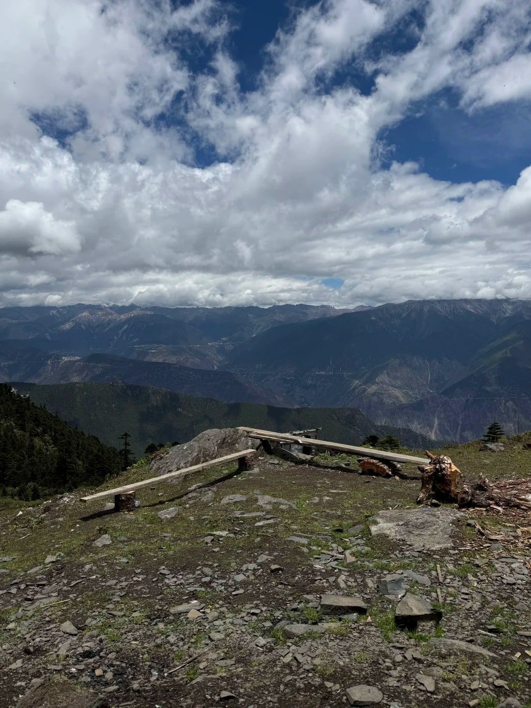 Zwei Bretterbänke auf einem steinigen Bergrücken, dahinter gestaffelte Bergketten und große weiße Wolken