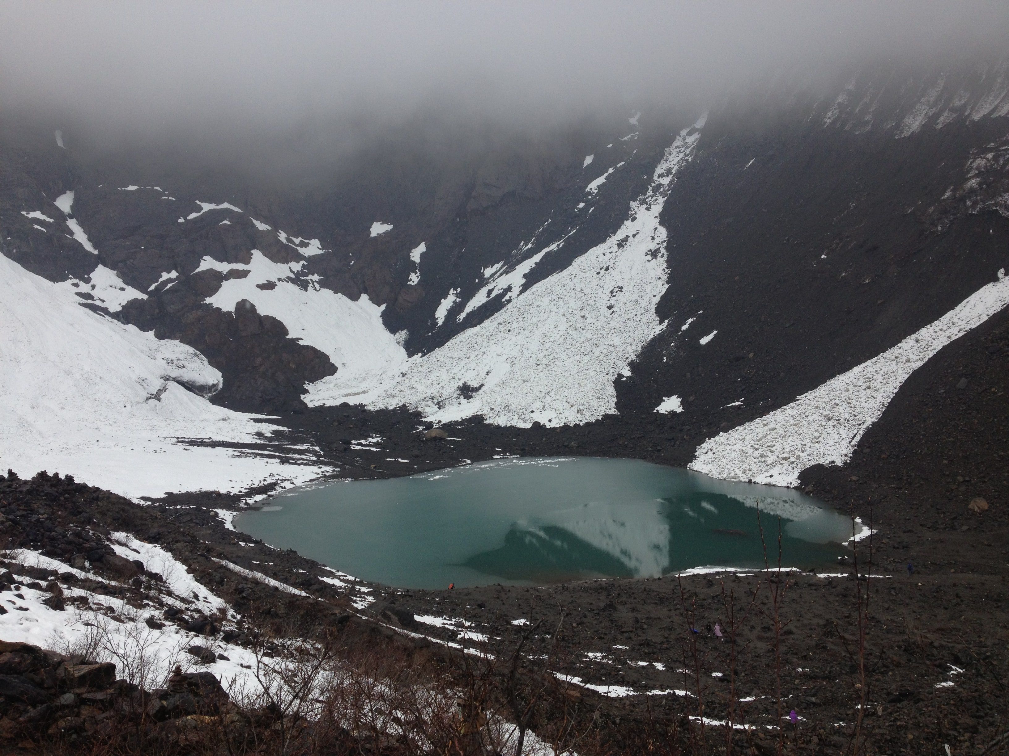 The green Ice Lake in a bowl of dark scree and old avalanche snow, cloud hiding the peaks above