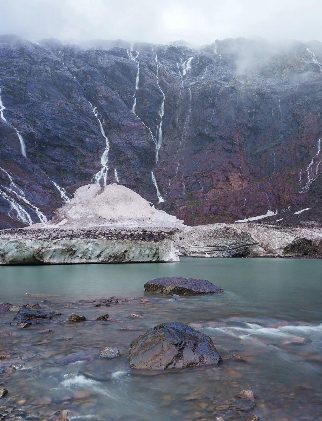 Ein milchig türkiser See unter dem Eisrand eines Gletschers und einer dunklen, von Wasserfällen durchzogenen Felswand an einem nebligen Tag