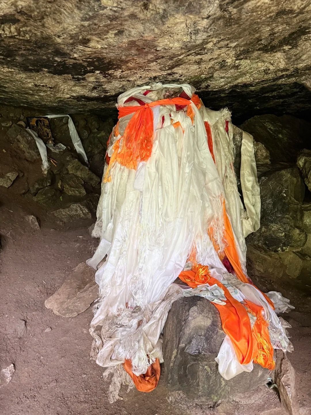 Ein Fels in einer niedrigen Höhle, behängt mit vielen weißen und orangefarbenen Khata-Schals