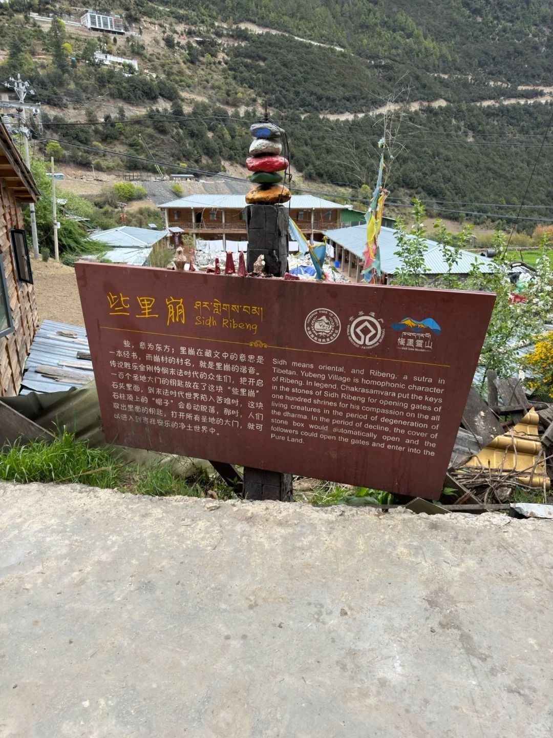 Die Tafel zu Sidh Ribeng auf Chinesisch, Tibetisch und Englisch, dahinter ein Pfosten mit bemalten Steinen und die Häuser des Dorfs