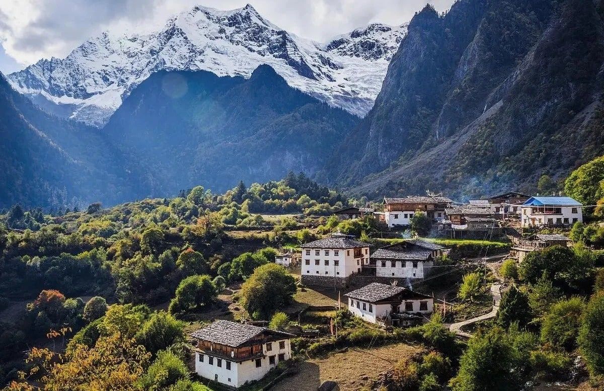 Weiß getünchte tibetische Häuser zwischen herbstlichen Bäumen in Ober-Yubeng, über dem Tal ein vergletscherter Schneegipfel