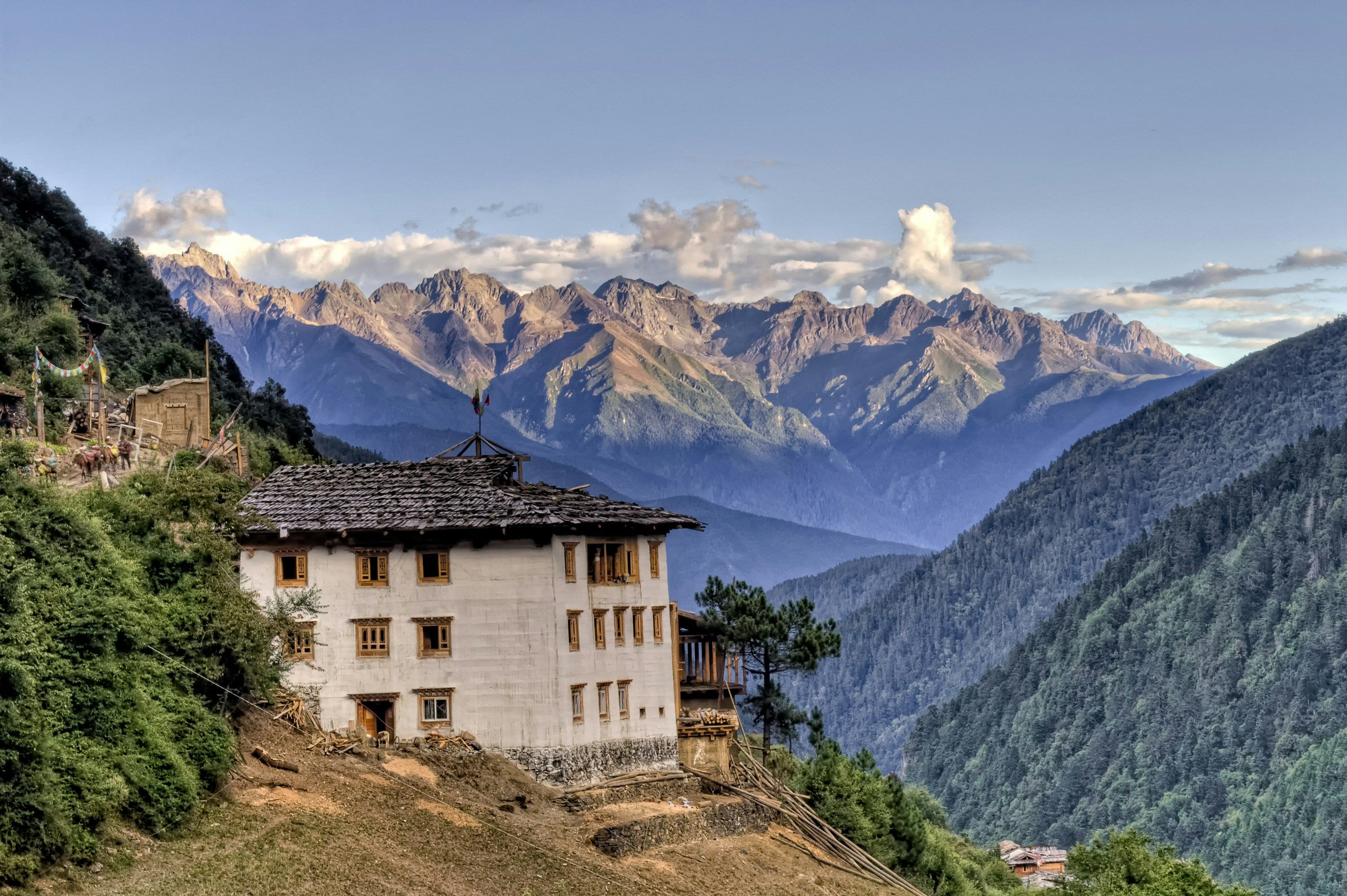 A whitewashed Tibetan house on a steep slope in Upper Yubeng, looking out over a forested valley to a long line of rocky peaks